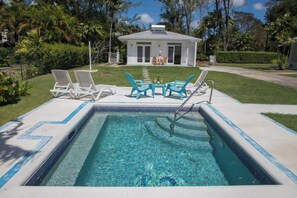 Une piscine rectangulaire dont le bord est recouvert de carreaux, entourée d’une terrasse en béton. On y trouve deux ensembles de chaises longues blanches et deux chaises turquoise. Une petite table est placée entre les chaises. Au fond, on aperçoit un bâtiment blanc doté d’un toit plat et d’un patio couvert.