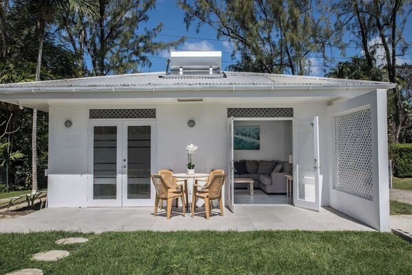 Un bâtiment blanc doté d’un patio couvert, d’un coin repas avec des chaises et une table, et d’un salon visible avec un canapé.