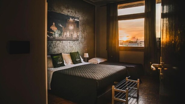 A room with a bed, a nightstand, a window with curtains, and a wall with a cityscape painting. - ALTA DIMORA b&b suites (Catania)