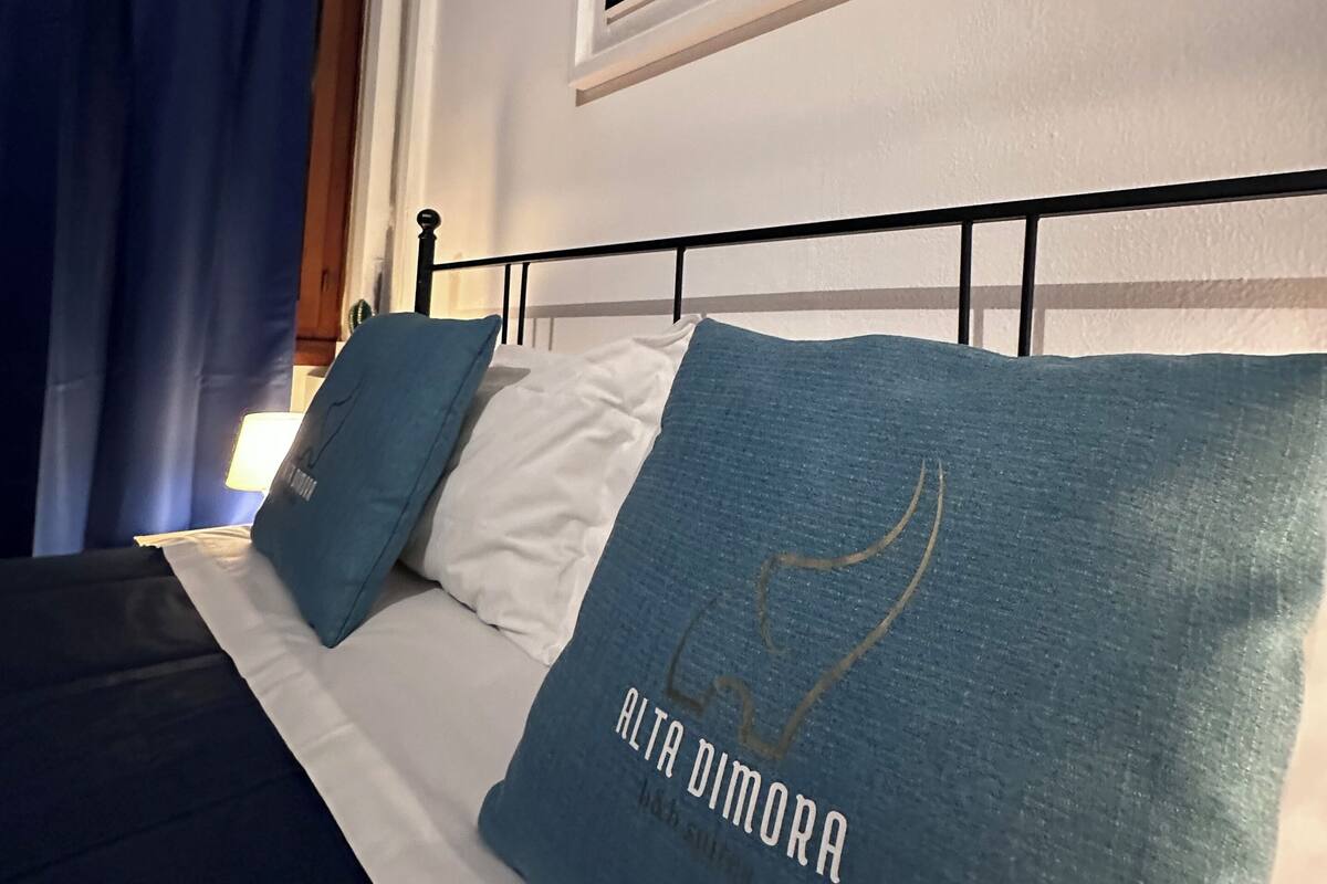 Ein Bett mit einer weiß-blauen Tagesdecke, zwei Kissen mit dem Aufdruck 'ALTA DIMORA', Blick auf einen Nachthimmel durch ein Fenster und ein blauer Vorhang.