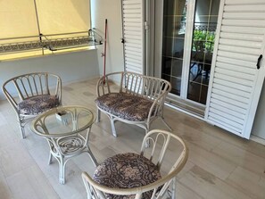 Een terras met vier rieten stoelen, een glazen tafel, een schuifpui en een raam met jaloezieën.