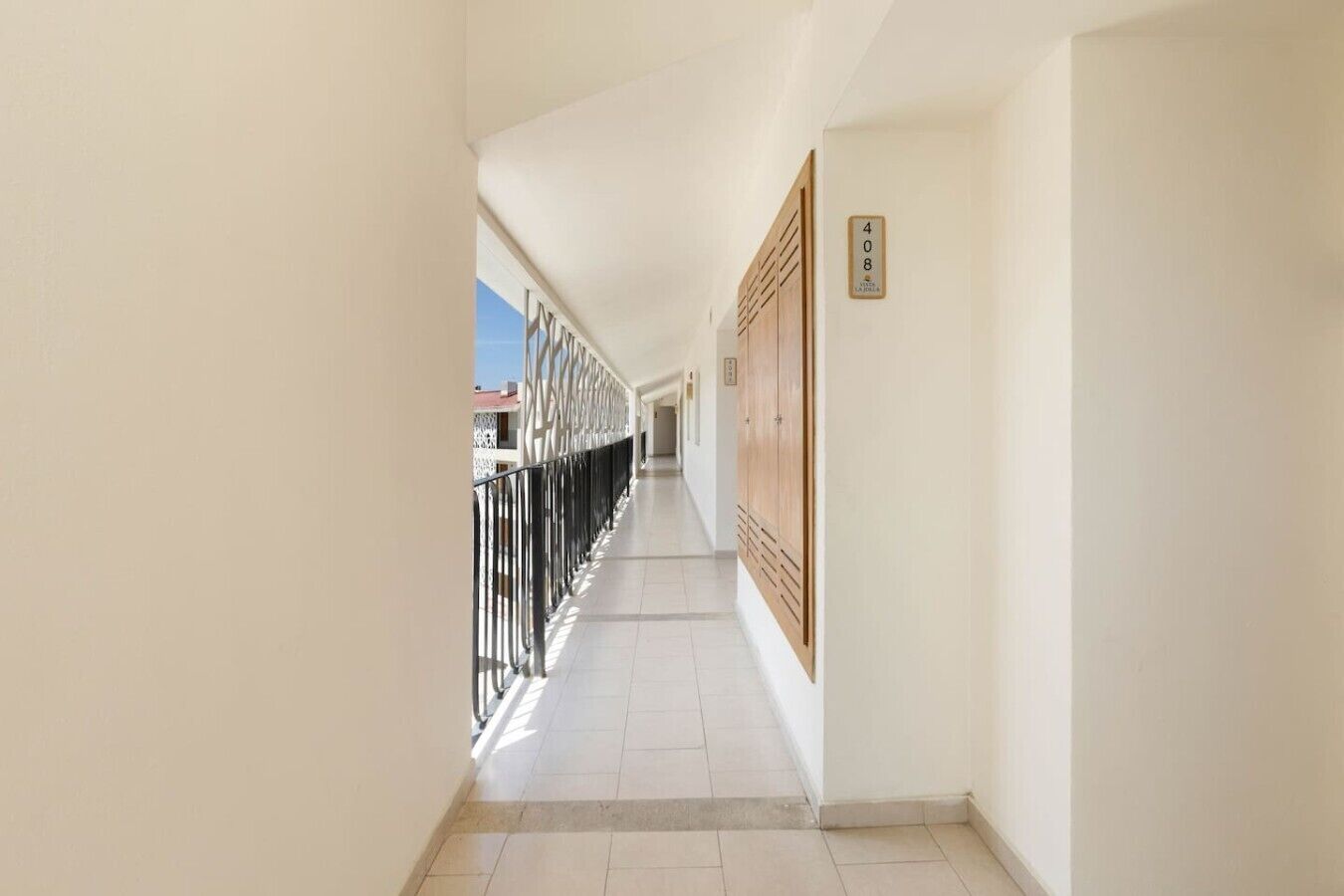 A corridor with a tiled floor, a wooden door with a number plate, and a metal railing on the left.