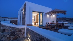 En moderne, hvid bygning med en stenvæg, en glasdørindgang og en patio med udendørs møbler og en stråtag parasol.