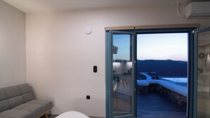 En moderne stue med en grå sofa, en hvid skammel og en glasdør, der fører ud til en balkon med udsigt over hav og bjerge.