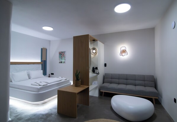 A modern bedroom with a bed, a sofa, a wooden side table, and a wall-mounted light fixture. - Thronos Suites (Mykonos)