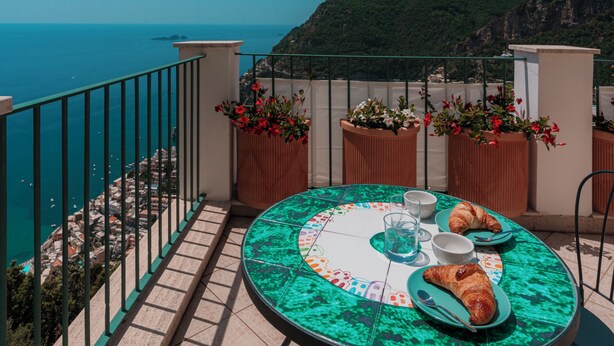 A round table with a green patterned tablecloth, two plates with croissants, and a glass of water on a balcony overlooking the sea. - Maison Ciccio - il Monticello with private Sauna (Campania)