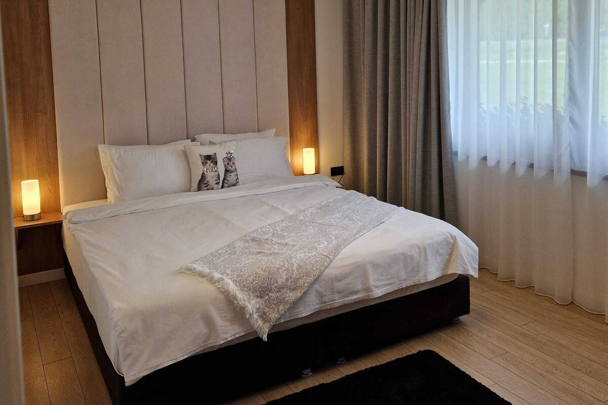 Une chambre moderne avec un grand lit, une tête de lit, une table de chevet, une lampe de chevet, un luminaire rond et des rideaux légers.