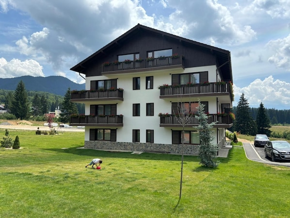 Un edificio de varios pisos con balcones, un cimiento de piedra y vistas a un campo verde y montañas al fondo.