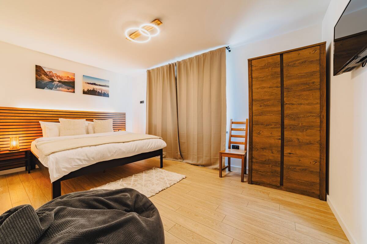 Une chambre moderne avec une tête de lit en bois, un lit recouvert de draps blancs, un plancher en bois, une grande fenêtre avec des rideaux et un téléviseur fixé au mur.
