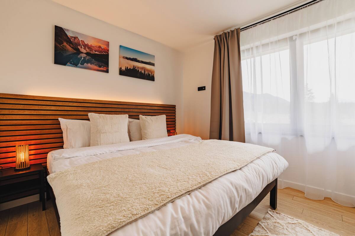 Une chambre moderne avec une tête de lit en bois, un grand lit recouvert de draps blancs, une couverture blanche et moelleuse, et deux tableaux représentant des paysages accrochés au mur.