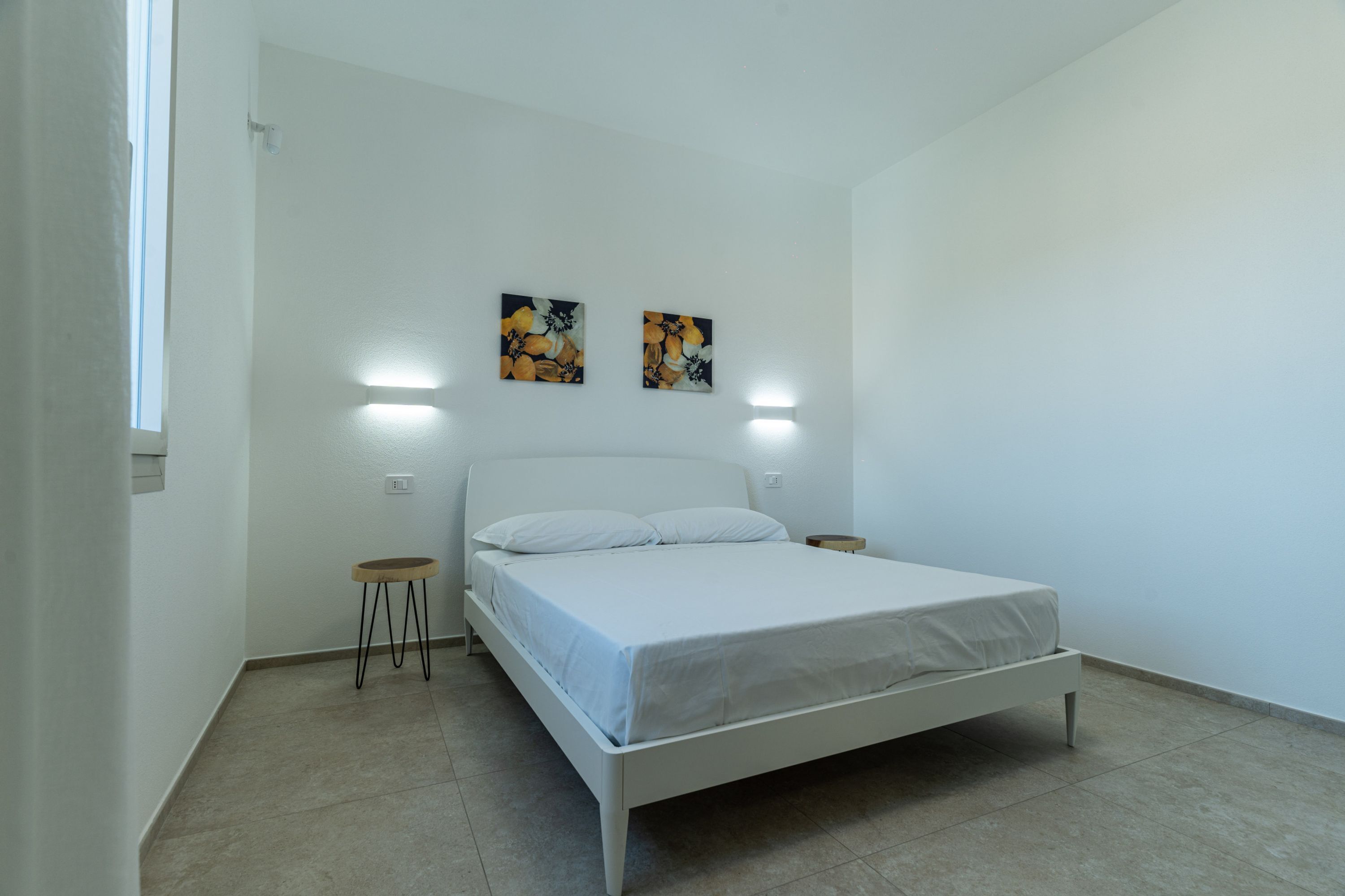 Une chambre avec un lit blanc, deux lampes fixées au mur et deux tableaux accrochés au mur.