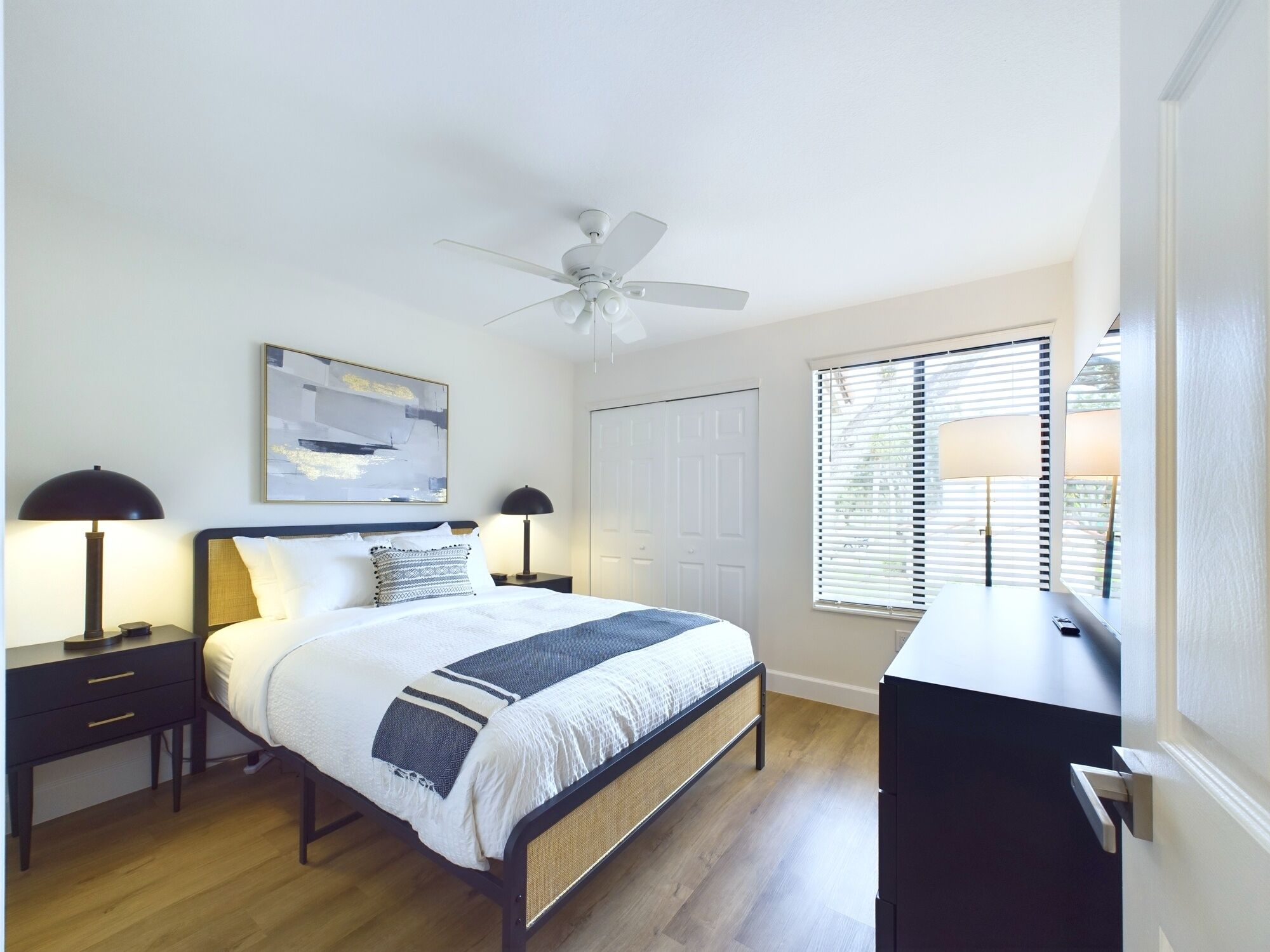 A bedroom with a bed, two bedside tables, a ceiling fan, a window with blinds, and a door.