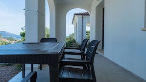 Een overdekt terras met een tafel en stoelen, witte muren en uitzicht op het landschap.
