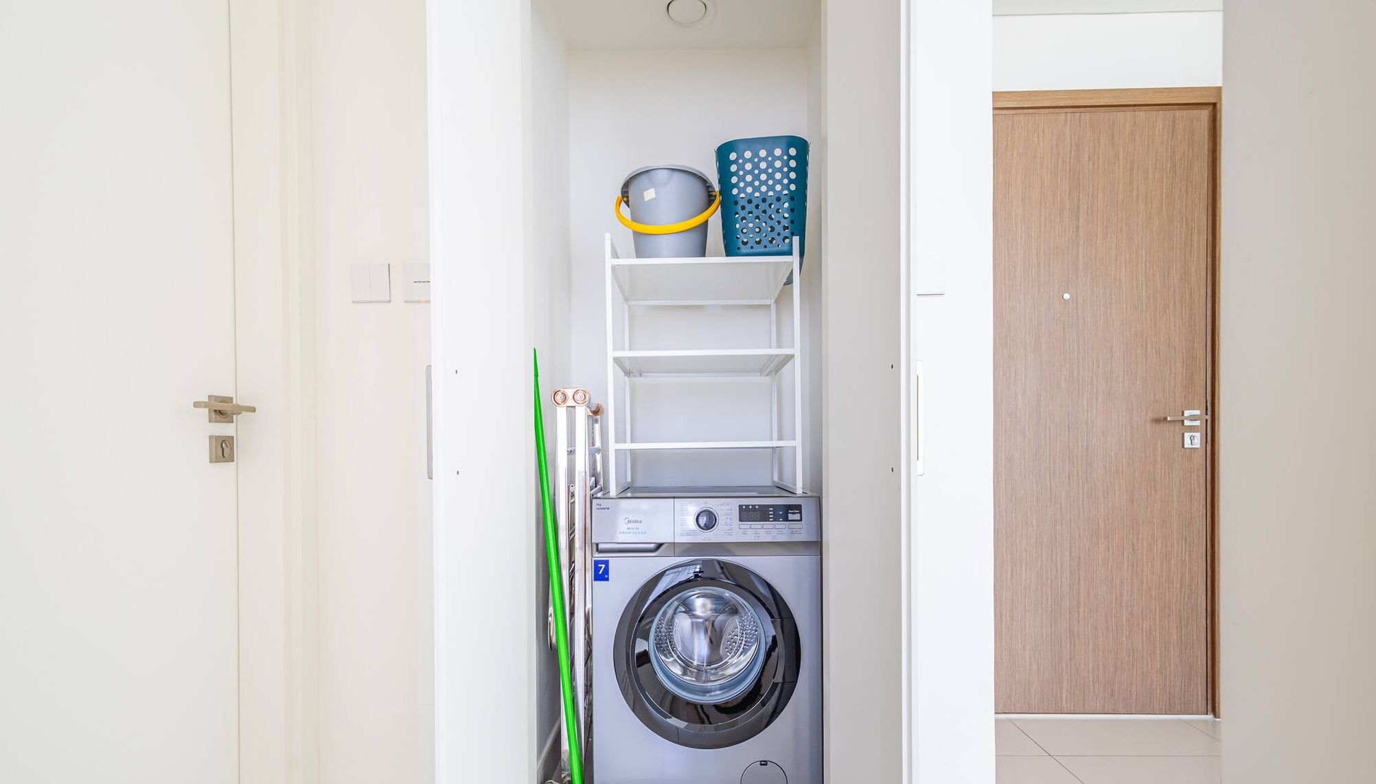 A compact laundry room with a front-loading washing machine, a drying rack, and a shelf with laundry baskets.