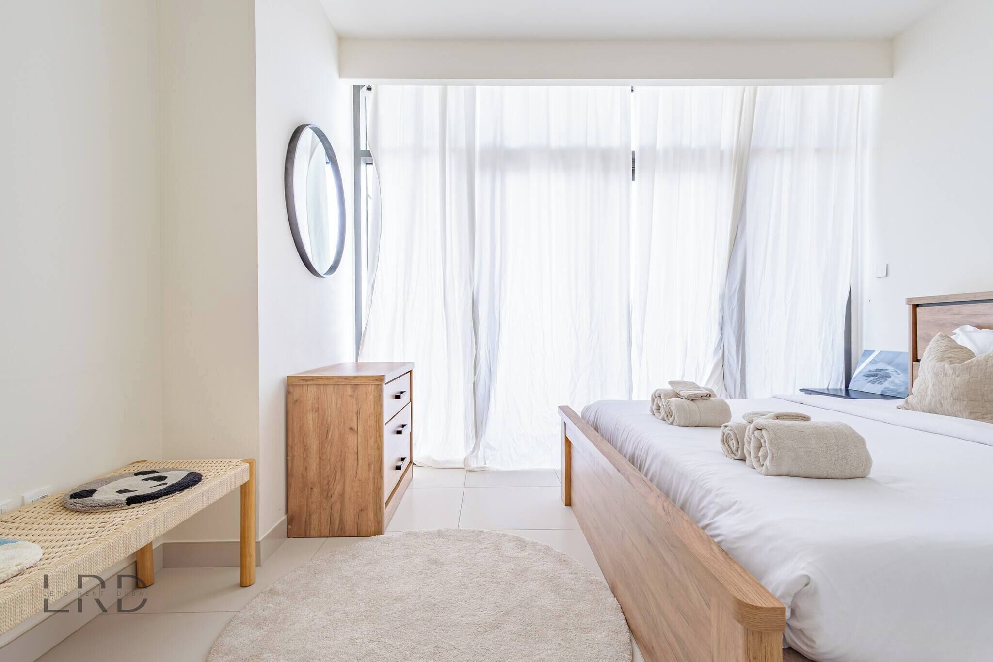 Een slaapkamer met een bed, een houten dressoir, een klein bankje, een ronde spiegel en dunne gordijnen.
