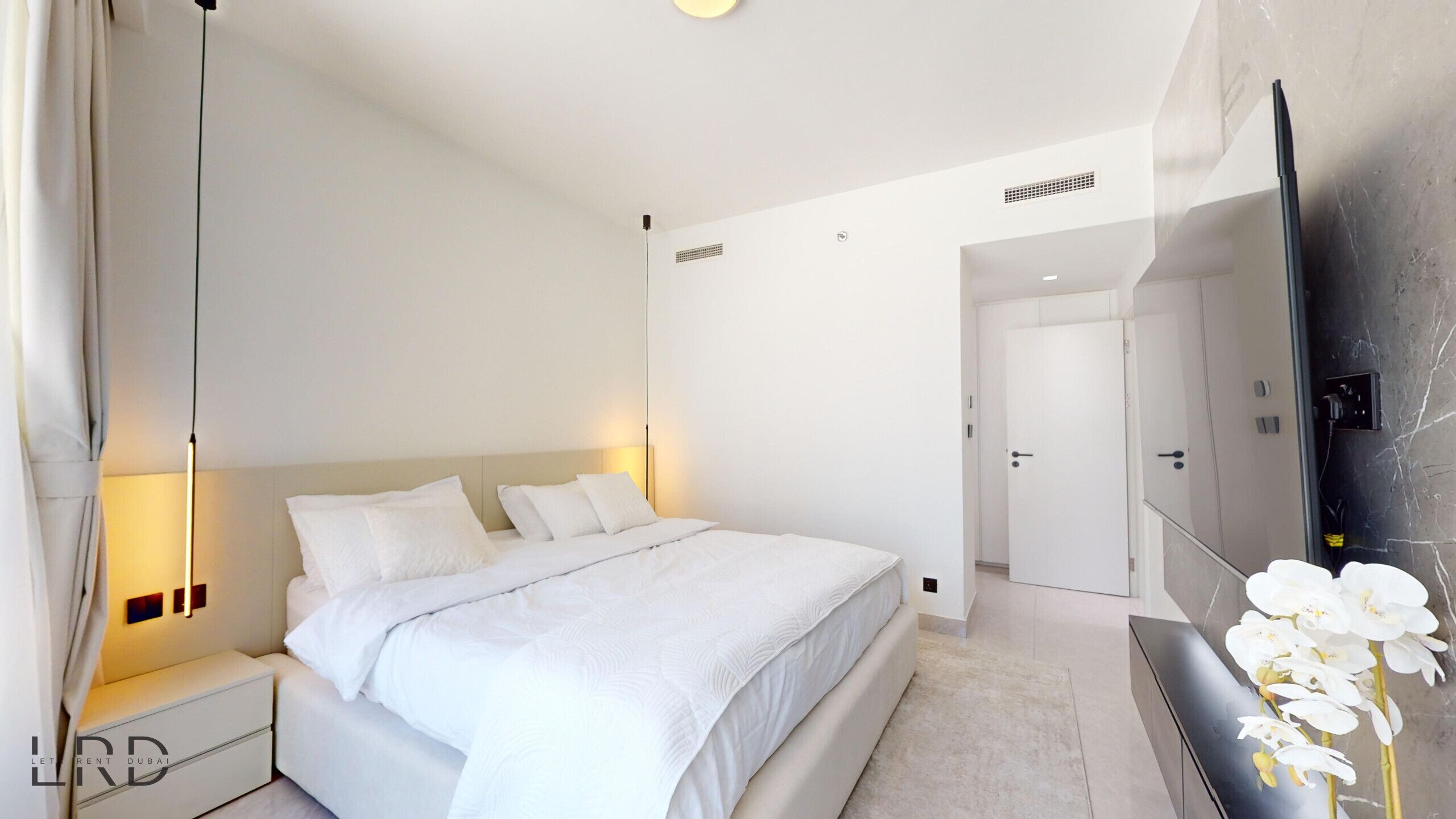 A modern hotel room with a large bed, bedside tables, a flat-screen TV, and a decorative orchid plant.