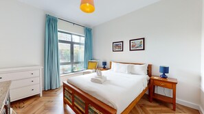A hotel room with a wooden bed, a white dresser, a nightstand with a lamp, and a window with curtains.