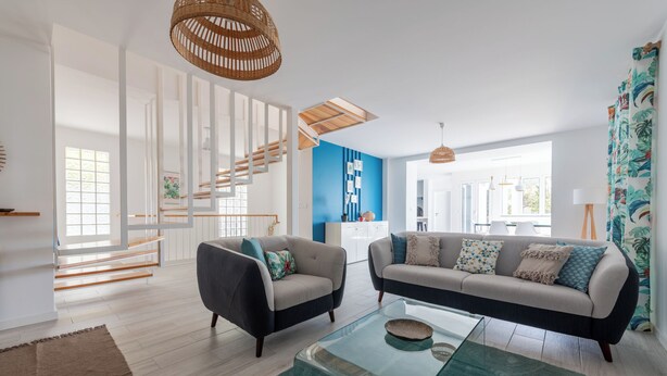A modern living room with a staircase, two sofas, a glass coffee table, and a kitchen area in the background.