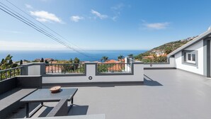 A rooftop terrace with a table and seating, overlooking a coastal landscape with houses and the sea.