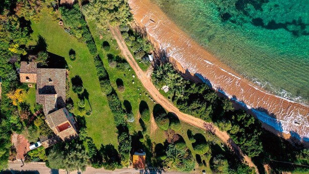 An aerial view of a coastal property with a beach, a fenced garden, and several buildings.