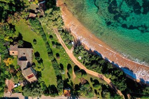 Vue aérienne d’une propriété située sur la côte, avec une plage, un jardin clos et plusieurs bâtiments.