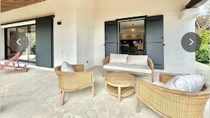 A modern outdoor seating area with wicker furniture, a ceiling with exposed wooden beams, and a sliding glass door leading to a dining area.