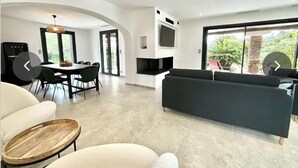 A modern living room with a dark sofa, a dining area with a table and chairs, and a large window with a view of greenery.