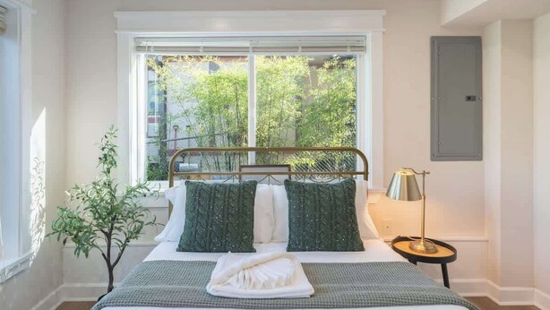 Un dormitorio con un ventanal grande, una cama con ropa de cama verde y blanca, una mesita de noche pequeña con una lámpara y una planta en maceta.