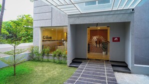 Entrada moderna de edifício com teto de vidro, pequena área ajardinada e sinalização com 'Liv' à esquerda.