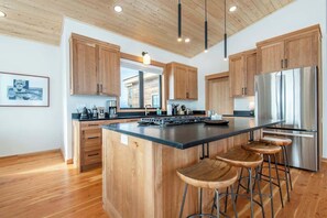 A modern kitchen with wooden cabinets, a large island with stools, stainless steel appliances, and a wooden ceiling.