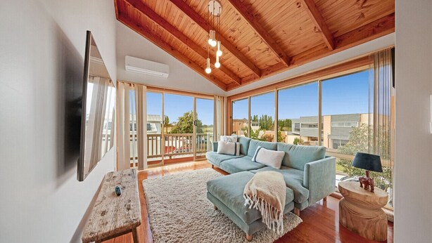 A spacious living room with a wooden ceiling, large windows, a sofa, and a TV mounted on the wall. - Sandy Bliss - Walk to the beach (Torquay)