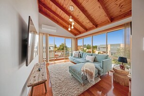 A spacious living room with a wooden ceiling, large windows, a sofa, and a TV mounted on the wall.