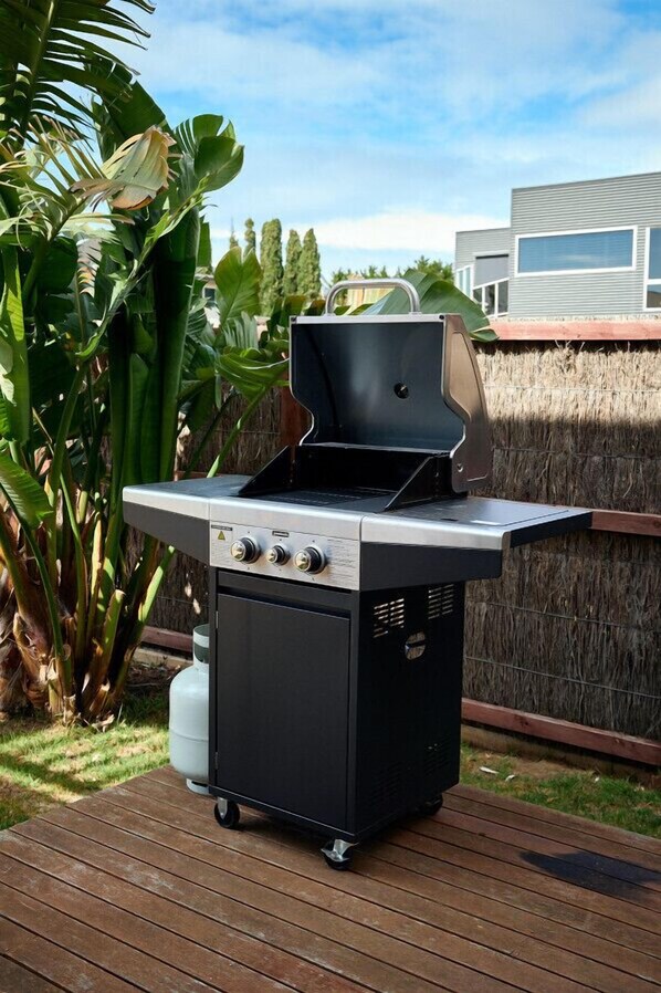 A portable grill with wheels, a control panel, and a gas tank, set on a wooden deck with a grassy area and a bamboo fence in the background.