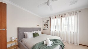 A bedroom with a bed, a ceiling fan, a window with curtains, and a wall-mounted artwork. - Sandy Bliss - Walk to the beach (Torquay)