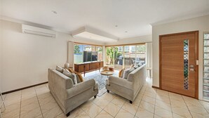 A spacious living room with two sofas, a coffee table, and a TV. There is a sliding glass door leading to a balcony with a view of the outside. - Sandy Bliss - Walk to the beach (Torquay)