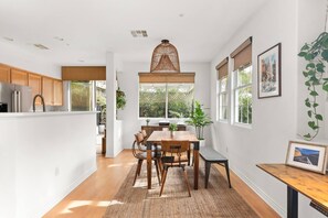 A modern kitchen with wooden cabinets, a dining area with a table and chairs, and large windows with blinds.