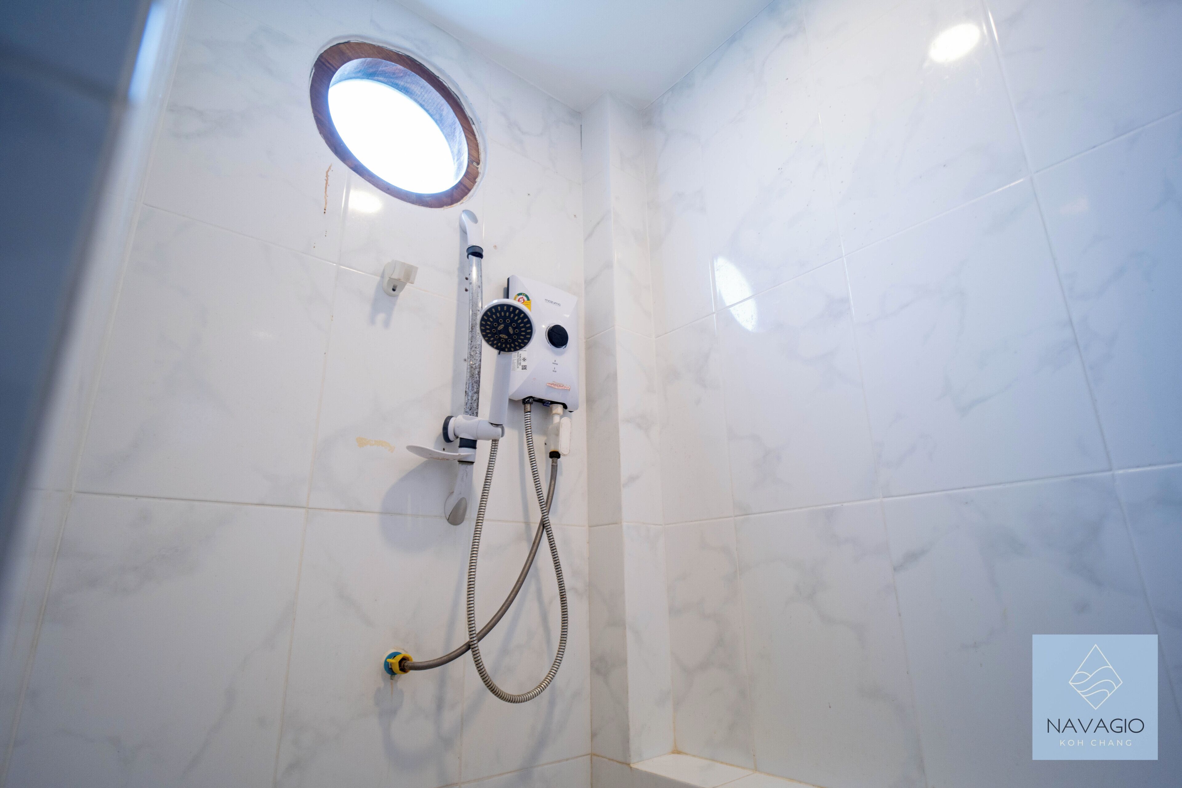 Un baño con azulejos de mármol blanco, ducha integrada con cabezal de ducha manual y una pequeña ventana circular.