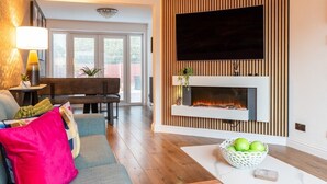 Una sala de estar moderna con chimenea, un televisor de pantalla plana montado en una pared de madera, y un comedor con mesa y sillas de madera.
