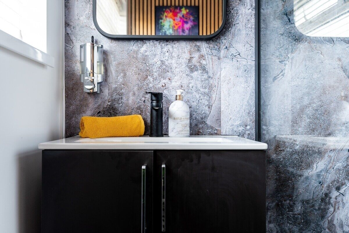 A bathroom with a marble wall, a black cabinet, a white sink, a yellow towel, a black bottle, and a mirror with a colorful artwork above it.