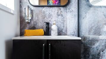 A bathroom with a marble wall, a black cabinet, a white sink, a yellow towel, a black bottle, and a mirror with a colorful artwork above it.