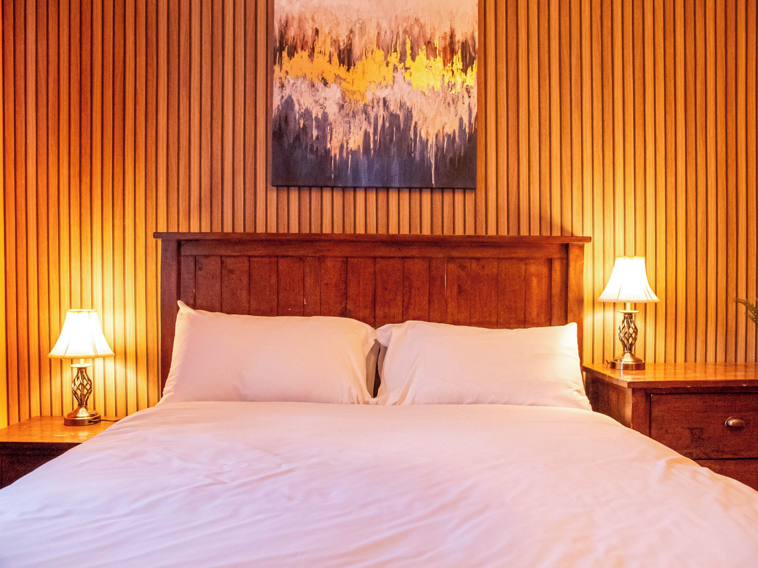 A hotel room with a wooden paneled wall, a large bed with white linens, two bedside lamps, and a painting above the bed.