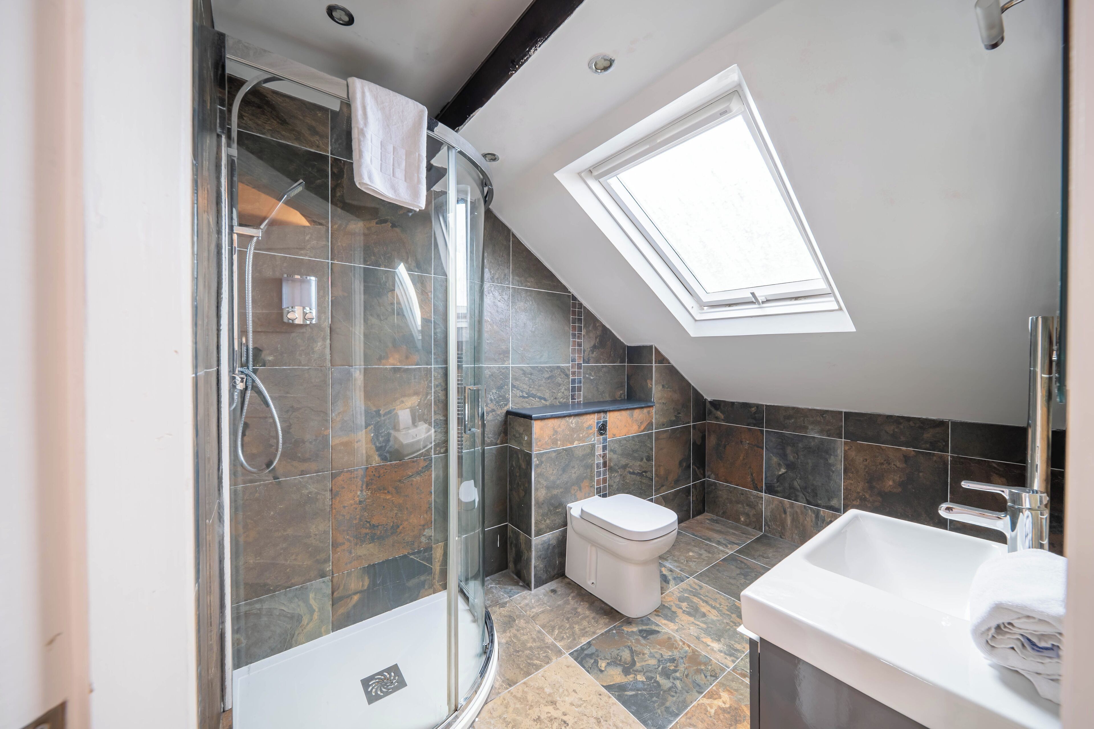 A modern bathroom with a glass-enclosed shower, a white toilet, and a skylight.
