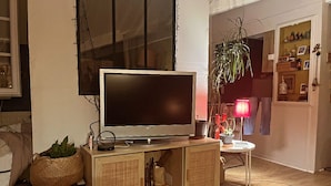 Una sala de estar con un televisor sobre un mueble de madera, una mesita con una lámpara y una planta en maceta.