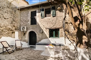 Un edificio de piedra con puerta negra, un pequeño porche con una mesa y dos sillones.