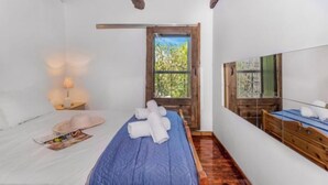 A bedroom with a wooden ceiling, a bed with a blue quilt, a nightstand with a lamp, a dresser, and a window with a view of trees. - Serene Retreat- Sleeps 4, Wifi, Courtyard (Lefkada)