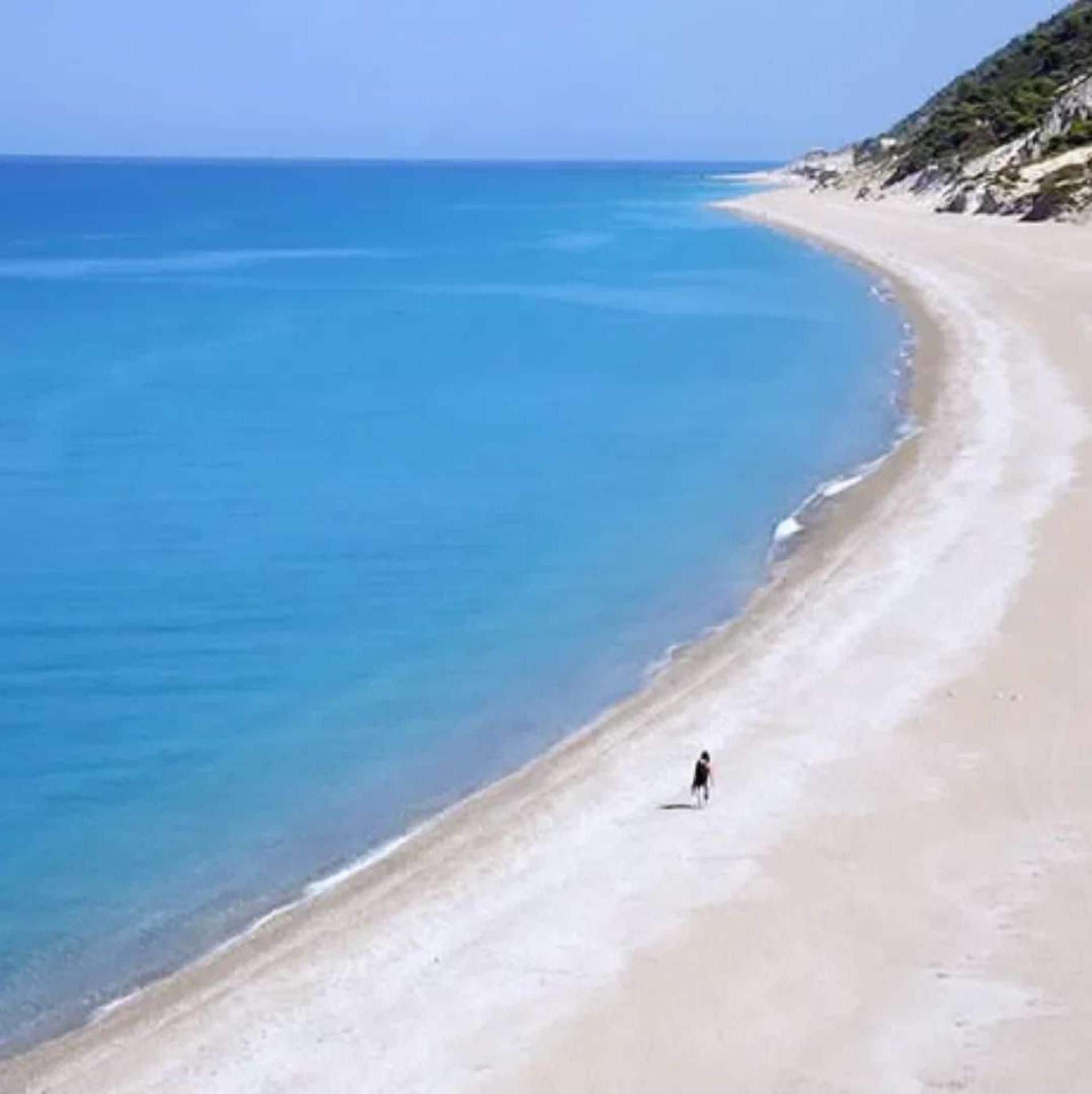 A beach with clear blue water, a sandy shore, and a person walking along the beach.