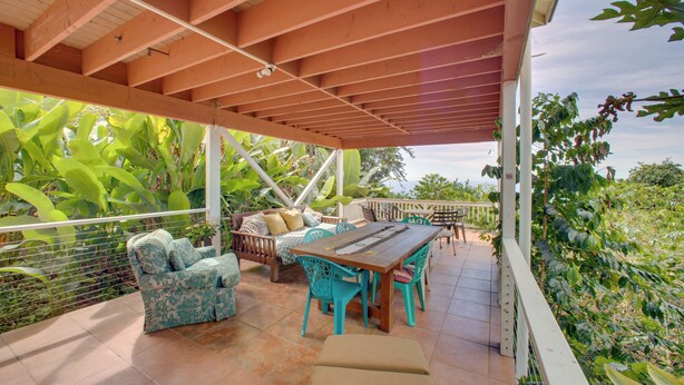 Ein überdachter Patio-Bereich mit Holztisch und -stühlen, einer Couch und einem Geländer mit Blick auf üppiges Grün.