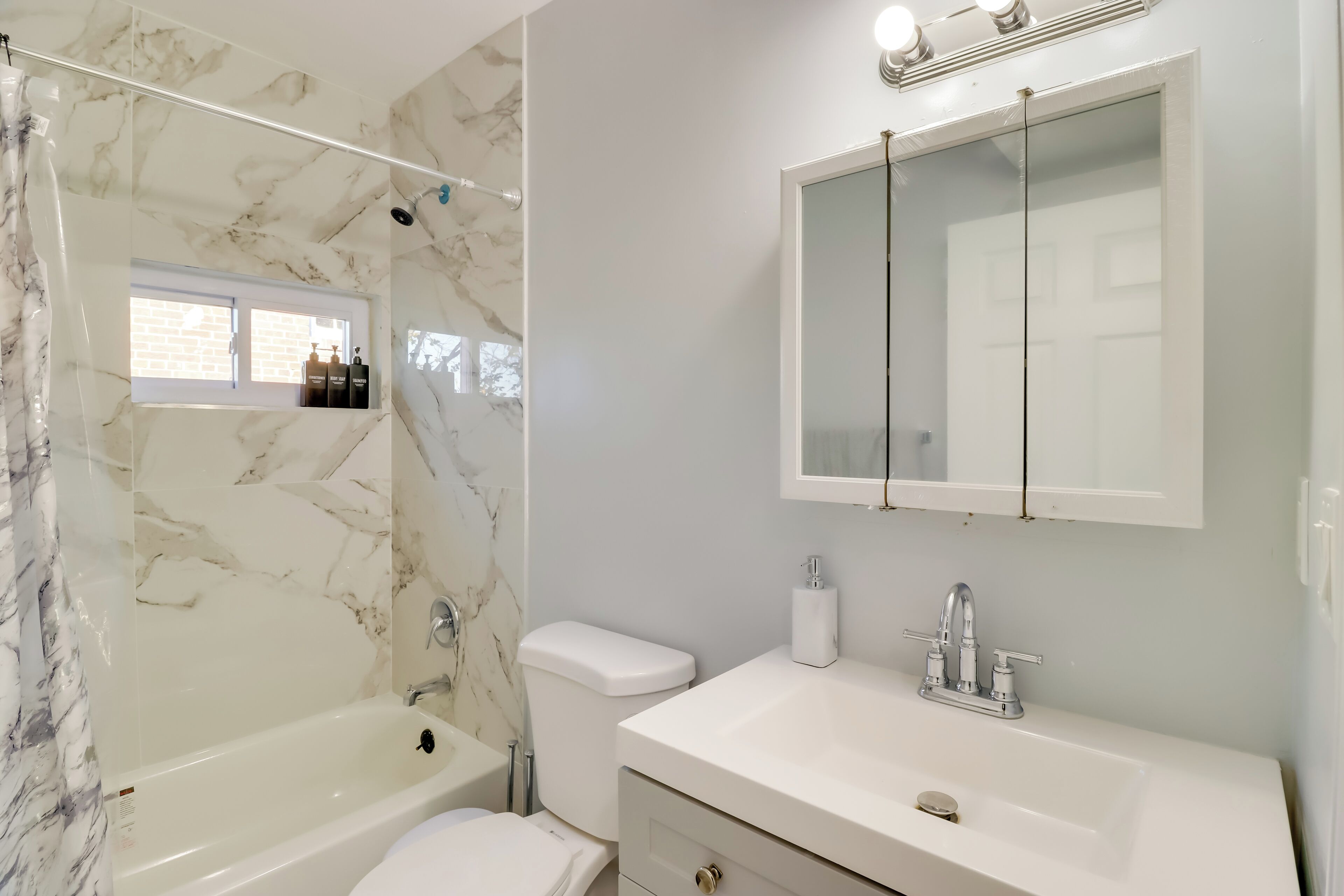 A bathroom with a marble shower, a toilet, a sink with a mirror, and a window with bottles on the shelf.