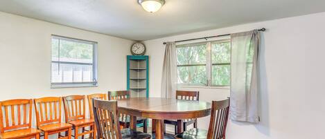 A dining area with a wooden table and chairs, a clock on the wall, and windows with curtains.
