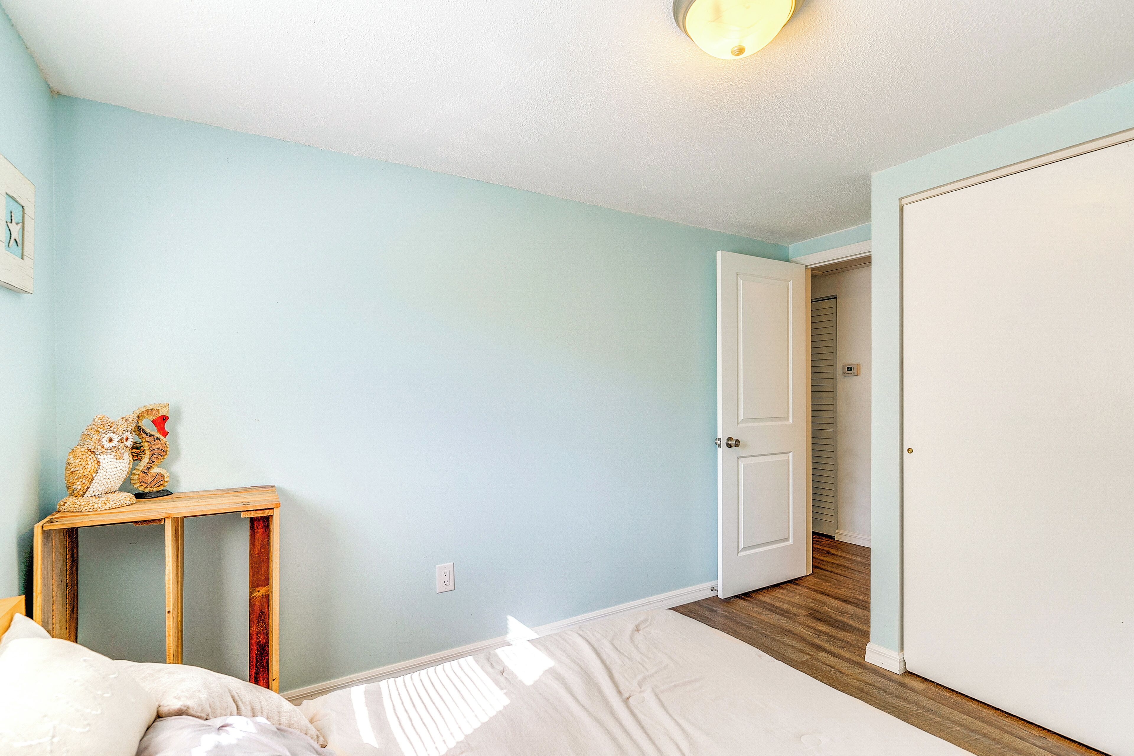 Un dormitori amb un llit, una tauleta de nit amb un auts decoratiu, una porta que condueix a una altra habitació i un interruptor de llum a la paret.